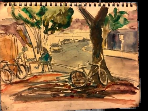 Watercolor sketch in a spiral notebook showing bicycles parked under a tree with buildings and street scene in background